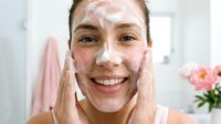 Young woman applying white facial cleanser foam while smiling in modern bathroom. Daily skincare routine, face washing, beauty treatment for healthy glowing skin.