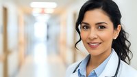 Young Hispanic female doctor smiling confidently in hospital corridor wearing white coat and stethoscope. Professional healthcare worker portrait.