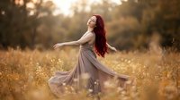 Young woman with long red hair dancing in flowing beige dress in golden autumn field at sunset with warm bokeh light creating dreamy romantic atmosphere.