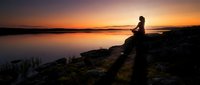 Woman sitting on rocky shore at sunset, meditating by calm water during golden hour. Peaceful solitude and nature contemplation.