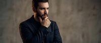 Thoughtful businessman in dark suit touching chin in contemplative pose against concrete wall background for corporate and professional concepts.