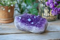 Raw amethyst crystal cluster on rustic wooden surface with potted flowers in background, showcasing natural purple mineral formation and crystalline structure.