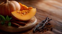 Fresh cut pumpkin slice with seeds on wooden board, whole orange gourd, vanilla pods, cinnamon sticks and mint leaves in warm autumn setting.