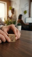Hand holding wooden cube with lightbulb icon on desk, representing innovation and creative ideas in modern office environment with colleagues working in background.