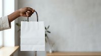 Man with dark skin tone holding blank white shopping bag with handles in bright interior space with plant and window light.