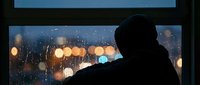 Silhouette of person looking through rain-covered window at blurred city lights creating bokeh effect during evening storm for contemplative mood concepts.