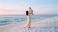 Young Caucasian mother in white dress lifting happy toddler on sandy beach at sunset, ocean waves and pastel sky in background.