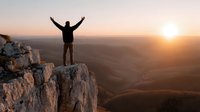 Silhouette of triumphant hiker with raised arms celebrating success on mountain cliff edge during golden sunrise over vast landscape valley below.