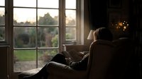 Young woman relaxing in cozy armchair by large window during golden hour sunset, peaceful home interior atmosphere for wellness and comfort concepts.