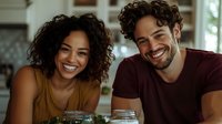 Happy young mixed race couple laughing together at kitchen table with mason jar drinks enjoying romantic morning date at home.