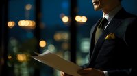 Professional businessman in elegant suit reading document against blurred city lights at night. Corporate executive reviewing paperwork in modern office setting.