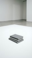 Stack of gray books on white floor in minimalist gallery space with concrete walls and modern interior design for contemporary art exhibition.