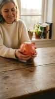 Smiling senior Caucasian woman holding a pink piggy bank at a wooden table, symbolizing retirement savings and financial planning.