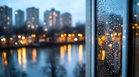 Rainy evening cityscape viewed through wet window glass with water drops, glowing lights reflecting in river, moody urban landscape with bokeh effect at dusk.