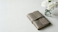 Elegant taupe leather journal tied with a strap beside white flowers in a glass vase on a light surface, minimal style.