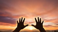 Silhouette of two hands reaching toward each other against vibrant sunset sky with orange and pink clouds creating dramatic atmospheric lighting.