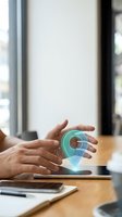 Hand holding glowing location pin hologram above wooden desk with tablet and smartphone, representing digital navigation, GPS technology, and mobile app development for business solutions.