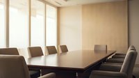 Modern corporate boardroom with large conference table and leather chairs in warm neutral office interior with natural window lighting.
