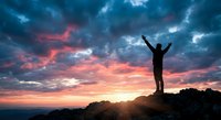 Silhouette of person with raised arms celebrating success on mountain peak against dramatic sunset sky with colorful clouds for motivation concepts.