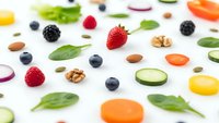 Colorful healthy food ingredients scattered on white background including fresh berries, nuts, vegetables and leafy greens for nutritious cooking.