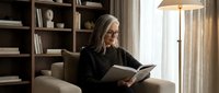 Middle-aged Caucasian woman with glasses reading book in cozy living room with bookshelves and warm lighting for relaxation and leisure concepts.