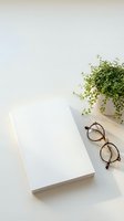 Clean minimalist workspace with blank notebook, eyeglasses and green plant on white desk for productivity and planning concepts.