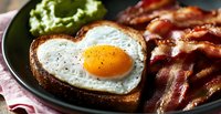 Heart shaped fried egg on toasted bread served with crispy bacon strips and fresh guacamole on dark plate, romantic breakfast close up.