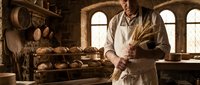 Professional baker holding wheat stalks in traditional stone bakery with fresh bread loaves and rustic ovens creating authentic artisanal atmosphere.