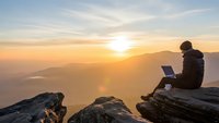 Digital nomad working on laptop at mountain summit during golden hour sunrise with layered peaks and warm sky creating inspiring remote work scene.