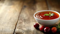 Fresh tomato soup garnished with basil leaf served in white ceramic bowl on rustic wooden table with cherry tomatoes and herbs, warm lighting.