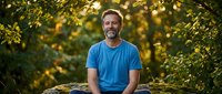 Middle-aged Caucasian man with beard wearing blue t-shirt sitting outdoors surrounded by golden foliage, suitable for lifestyle, wellness, and portrait projects.