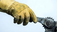 Worker's hand in worn yellow leather glove gripping a metal wrench, close-up shot emphasizing industrial labor and manual work safety.
