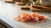 Thinly sliced cured ham arranged on a marble surface with warm sunlight, wooden boards and olive branch in the background.