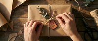 Hands wrapping kraft paper gift box with twine and wooden tag on rustic wooden table, overhead view for holiday packaging and present preparation projects.