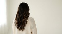 Young woman with long wavy brown hair wearing cream sweater standing by window in minimalist room, back view lifestyle portrait for wellness concepts.