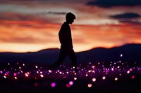 Silhouette of young man walking through magical field of glowing purple lights against dramatic orange sunset sky and mountain landscape. Dreamy atmosphere.