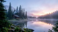 Serene mountain lake at dawn with wooden cabin among evergreen trees, misty water reflections and soft purple pink sunrise sky creating peaceful wilderness atmosphere.