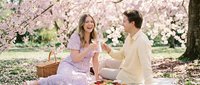 Young couple enjoying spring picnic under blooming cherry blossom trees, sharing food and romantic moments outdoors during springtime celebration.
