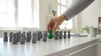 A hand with medium skin tone reaches toward a green peg among gray pegs on a white table, symbolizing recruitment, selection, and business strategy for corporate hiring concepts.