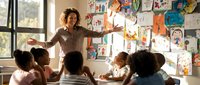 Smiling female teacher with curly hair engages diverse group of young students in bright elementary classroom with colorful artwork on walls.