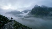 Solitary hiker stands on mountain ridge overlooking misty valley with dramatic fog-covered peaks, capturing adventure and exploration in wilderness landscape.