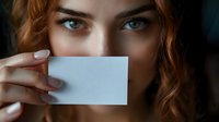 Close-up portrait of young woman with red hair and blue eyes holding blank business card over mouth against dark background.