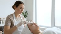 Professional female cosmetologist performing facial treatment on relaxed client in modern spa salon with natural lighting for skincare wellness.