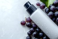 White cosmetic bottle with pump dispenser surrounded by fresh purple grapes on textured surface. Natural skincare product mockup for beauty and wellness branding.