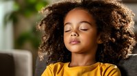 Young African American girl with curly natural hair sleeping peacefully in yellow t-shirt, head tilted back on sofa, soft natural lighting creates serene atmosphere.