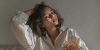Young woman with curly hair in white shirt posing gracefully with hand in hair, soft natural lighting for lifestyle and beauty content.