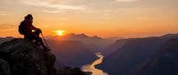 Hiker sitting on mountain peak at sunrise overlooking winding river valley below, contemplating scenic landscape during golden hour adventure and exploration.