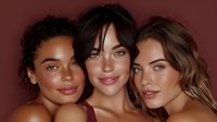 Three diverse young women with glowing skin posing together against burgundy background for beauty and skincare advertising campaigns.