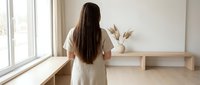 Young woman with long dark hair in white shirt standing by window in minimalist modern interior with wooden furniture and pampas grass decoration.