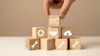 24/7 customer service concept with hand placing wooden block featuring service icon above stacked cubes displaying support, gear, wrench, and cloud symbols on neutral background.